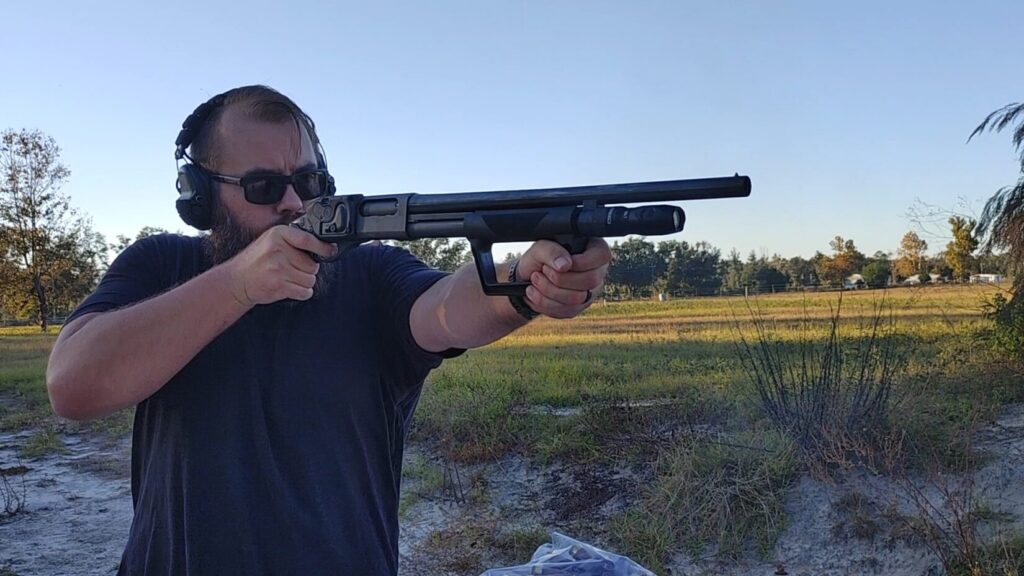Man with beard and sunglasses shooting a short-barreled shotgun.