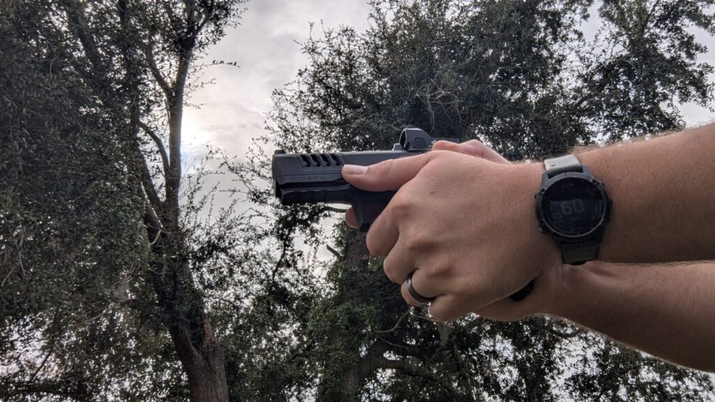  Shooter in a two-handed grip aiming a pistol with a red dot sight against a backdrop of trees and a cloudy sky.