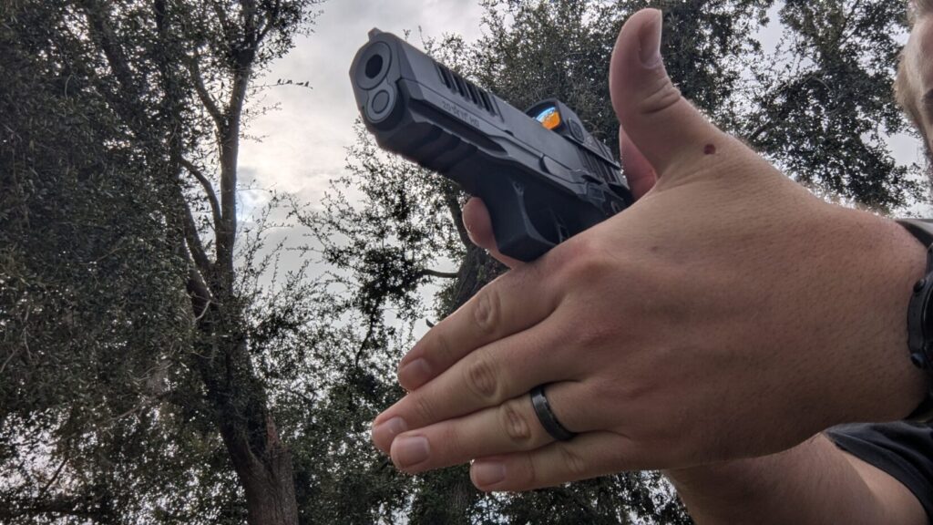  Shooter in a two-handed grip aiming a pistol with a red dot sight against a backdrop of trees and a cloudy sky.