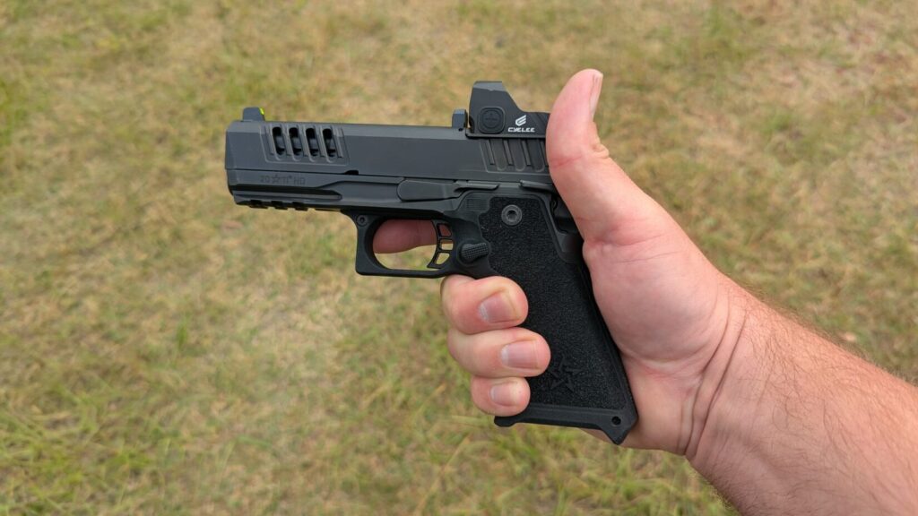 A hand holding a black competition-style pistol with a ported slide and a mounted Cyelee red dot sight over a grassy background.