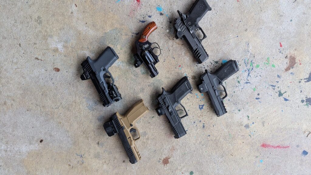 Overhead view of a collection of six pistols and a revolver lying on a colorful concrete background.