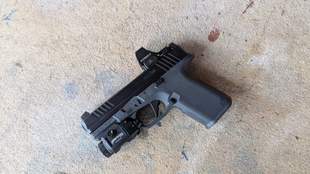 Gray and black pistol with a red dot sight and a weapon light, lying on concrete.
