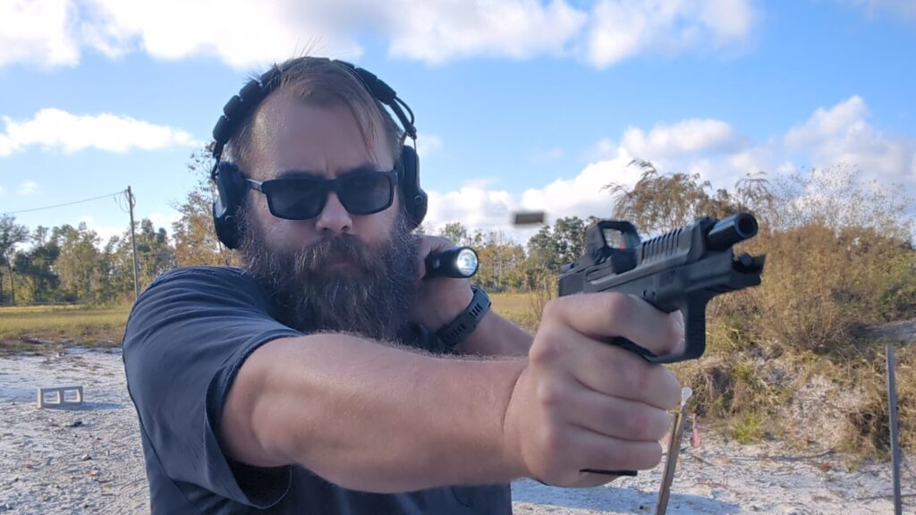 Man aiming pistol outdoors, holding a flashlight in a compressed grip against his neck