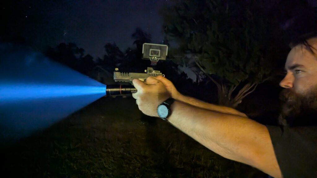 Close-up of a man using the Rogers/SureFire technique, aiming a pistol and handheld flashlight outdoors at night.