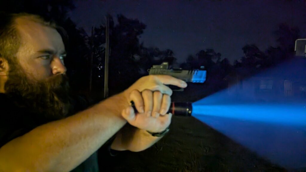 Profile of a man using the Harries technique, aiming a pistol and shining a bright handheld flashlight in the dark.