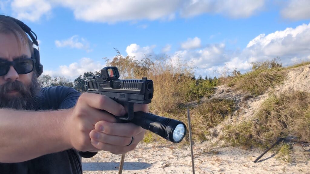 Man aiming pistol with a large handheld flashlight braced against the firearm outdoors Surefire Technique
