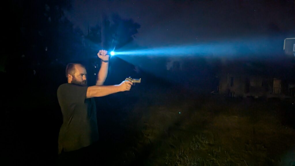 Man aiming a pistol outdoors at night, holding a flashlight overhead with a bright beam.