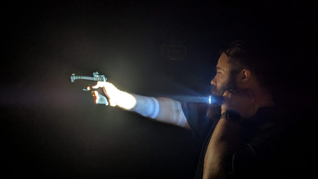 Man in the dark aiming a pistol with a weapon light attached, the beam illuminating his arm.
