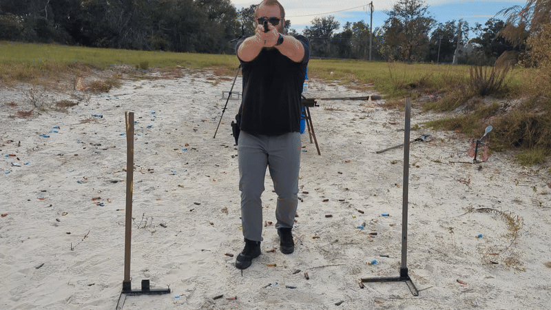Man walking backward while aiming handgun past a blue barrel.