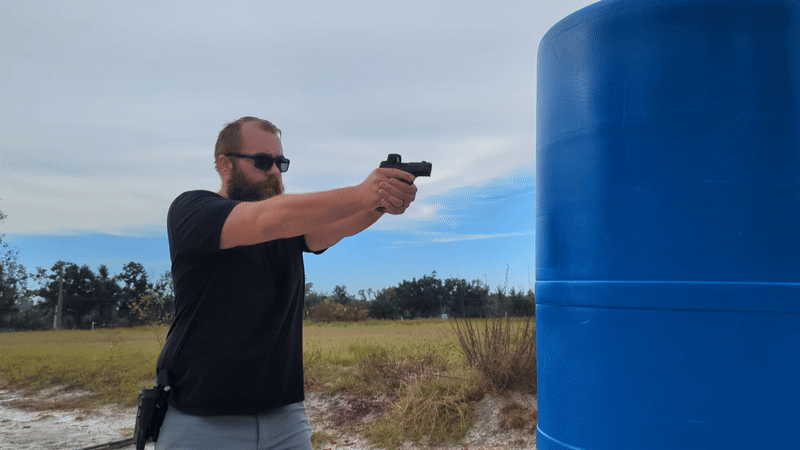 Man aiming handgun past a large blue barrel on a sunny range.