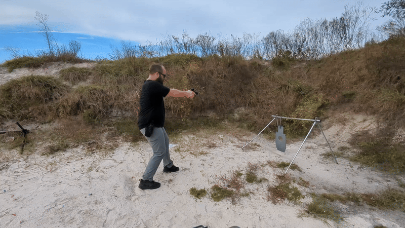 Man shooting handgun at a steel target on an outdoor range.