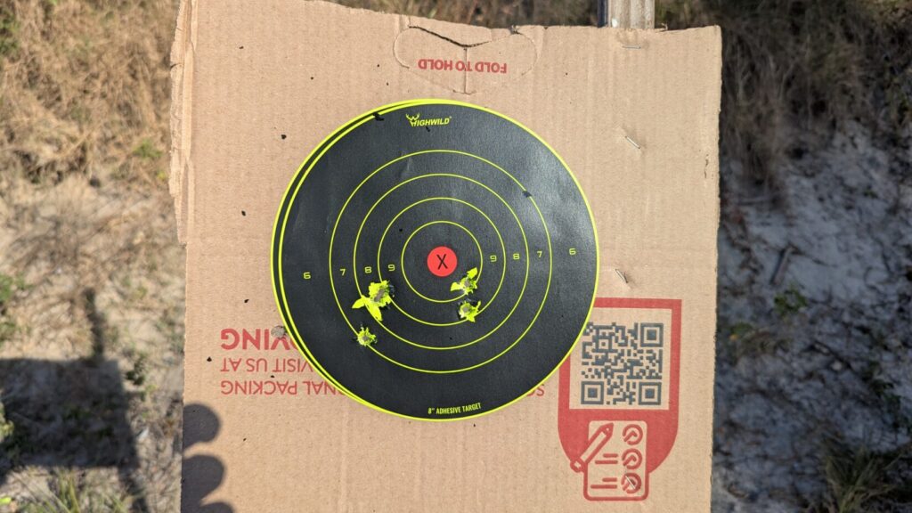 Circular shooting target with a yellow ring and bullet holes in the lower left quadrant on a cardboard backer.