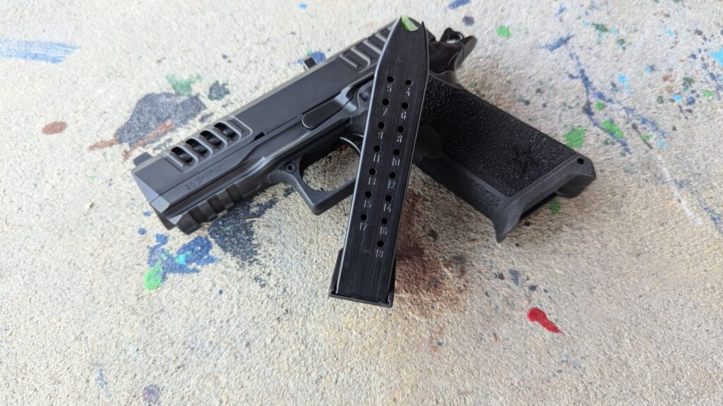 A long, double-stack pistol magazine (showing '18' capacity mark) resting next to the Staccato HD P4 on a paint-splattered concrete surface.
