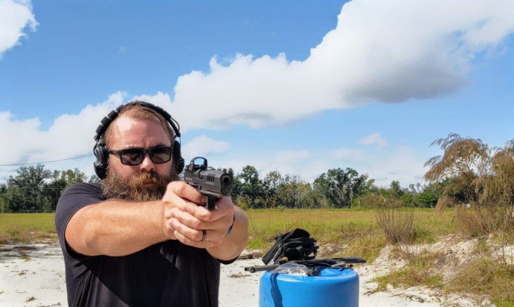 Man with beard, sunglasses, and hearing protection aims a Staccato HD pistol with a red dot sight straight ahead at an outdoor range.