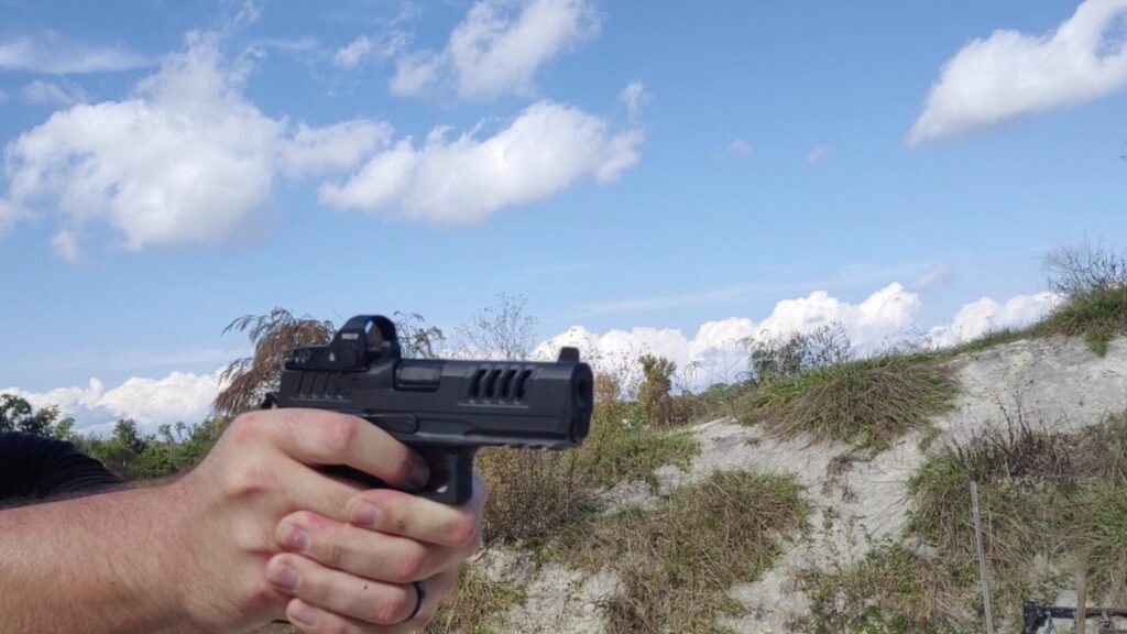 Close-up of a hand holding a Staccato HD pistol with slide cuts and a red dot sight, aimed toward a grassy hill under a cloudy blue sky.