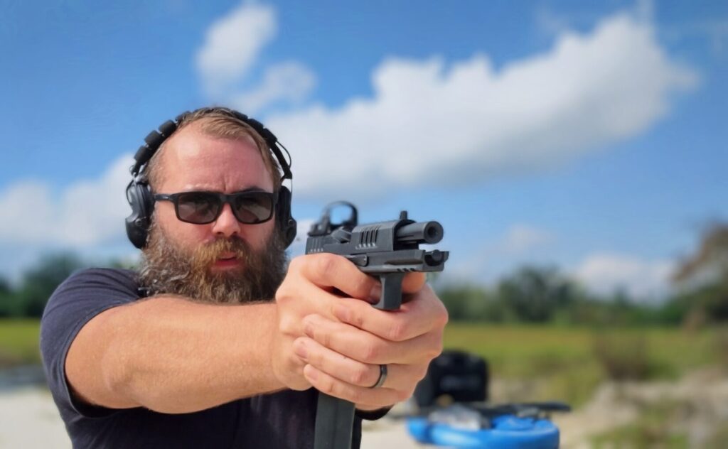 man shooting a Staccato HD pistol with slide cuts and a red dot sight, aimed toward a grassy hill under a cloudy blue sky.