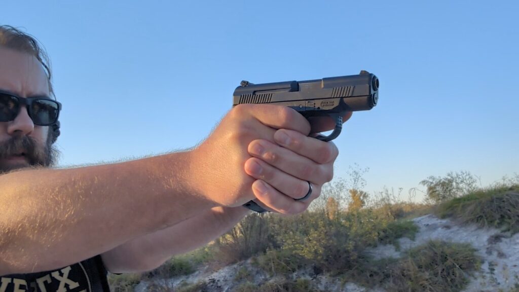 Man with beard and sunglasses aiming the SW99 pistol outdoors.