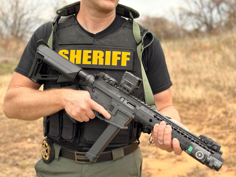 A person in a black vest with yellow "Sheriff" on the vest, holding an AR-15 style rifle. 