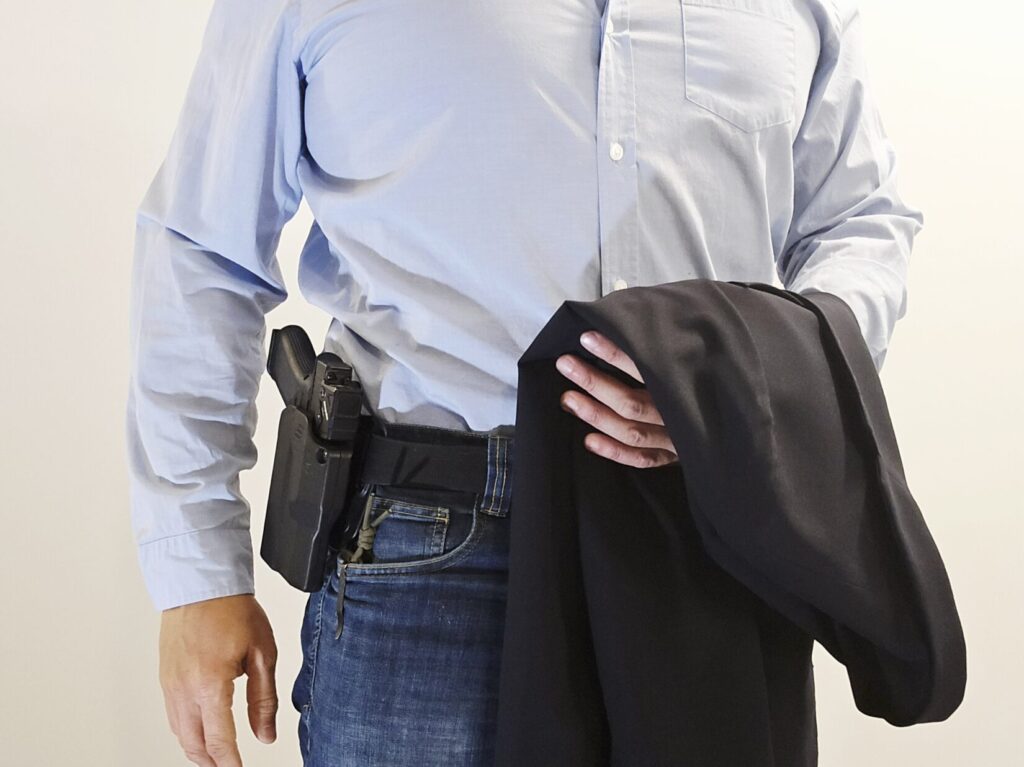 A man in a blue shirt and jeans holding a sports coat with a pistol on his belt
