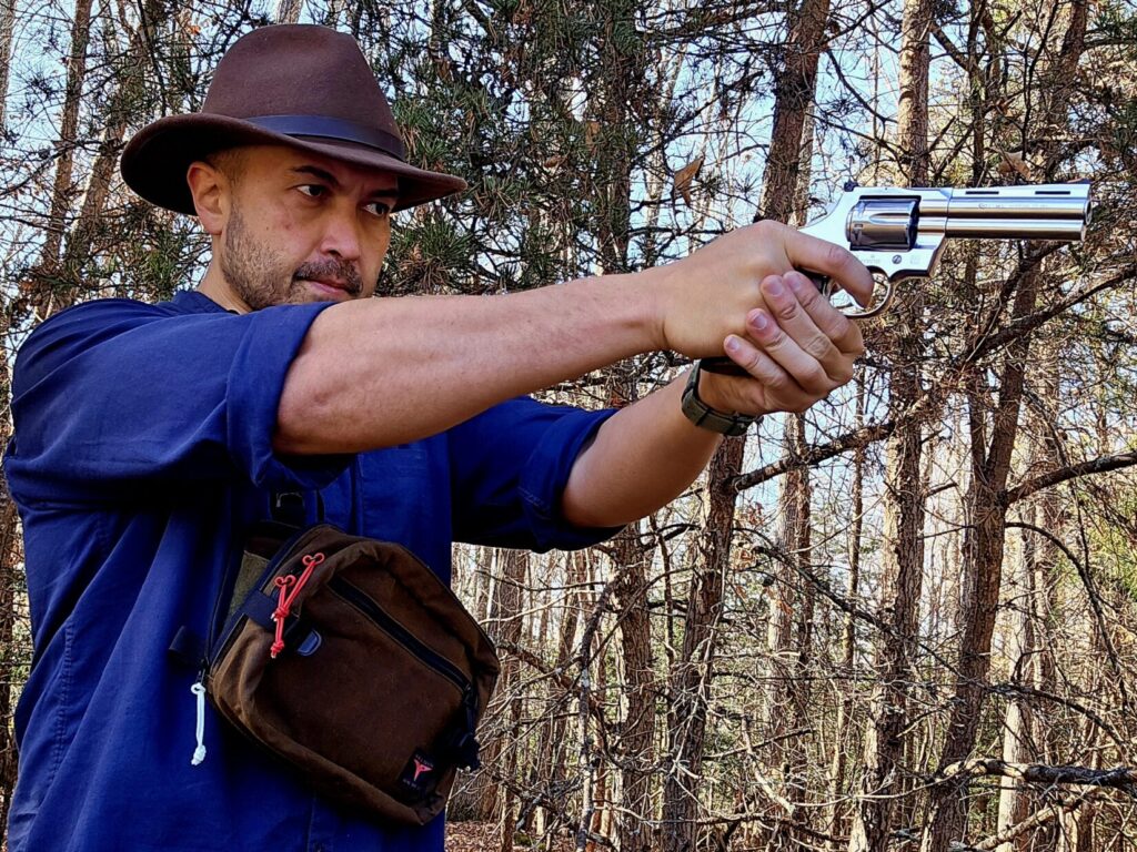 A man in a blue shirt and brown hat takes aim with a stainless revolver in a wooded environment