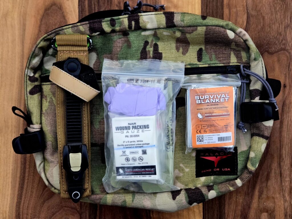 A tourniquet medical pouch and survival blanket on top of a multicam pack on a wooden backdrop