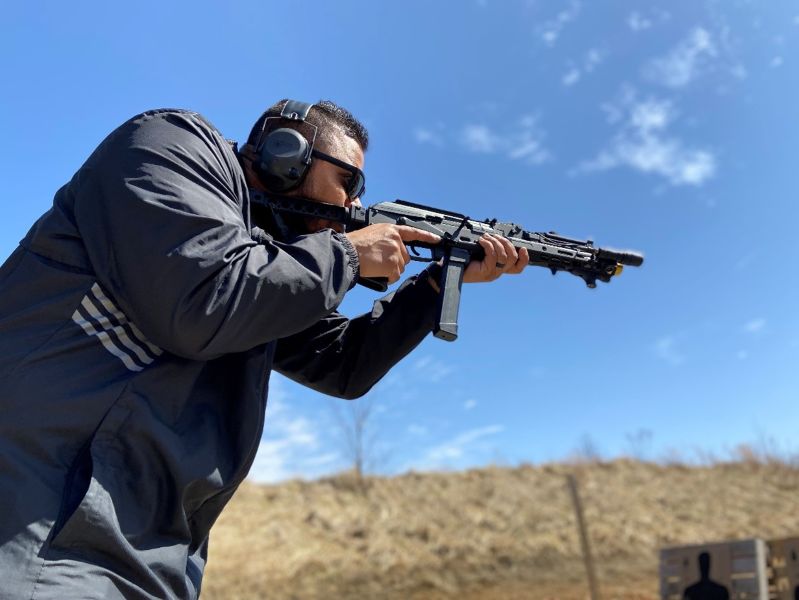 A person in a blue jacket, shooting an AK style rifle with a Glock magazine in it on a shooting range.