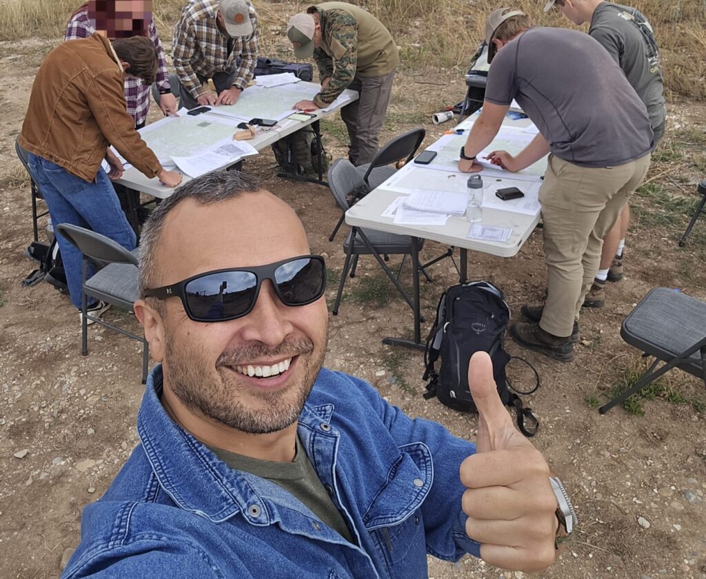 A man in a blue denim shirt with people working with maps and compasses in the background