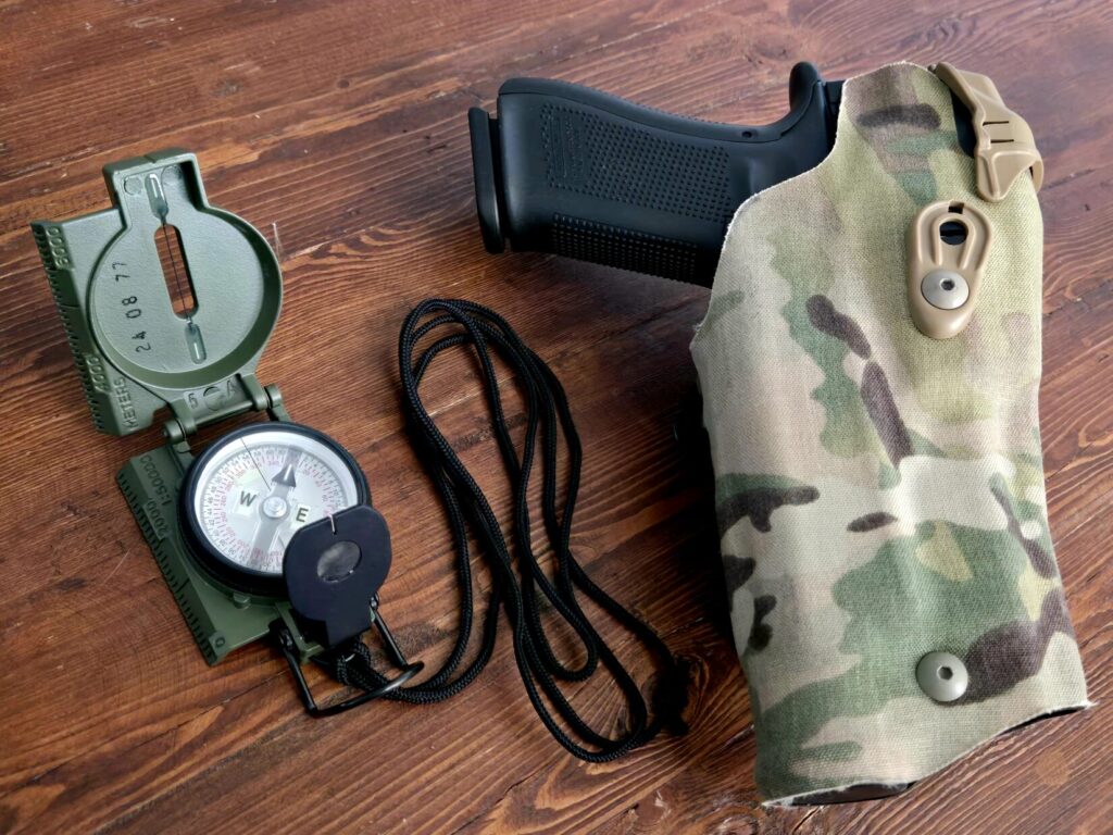 A military lensatic compass next to a multicam holster for a glock pistol