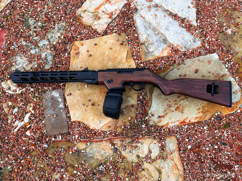 A wood rifle with a black drum and black barrel, laying on some tan rocks. 