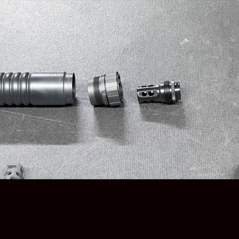 A silencer on a grey table with a mount next to it, and a muzzle device next to that, showing how a silencer attaches to a rifle. 