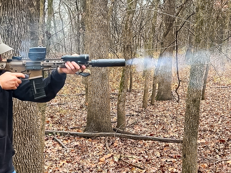 A person shooting a tan rifle in the woods. Smoke is coming from the end of the silencer. 