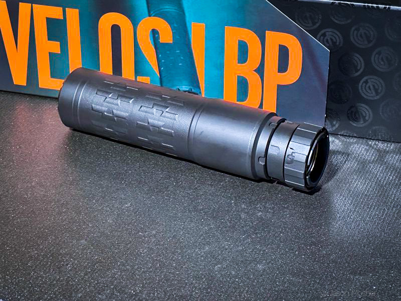 A black silencer on a black surface with a box behind it, displaying the "VELOS" name in orange. 
