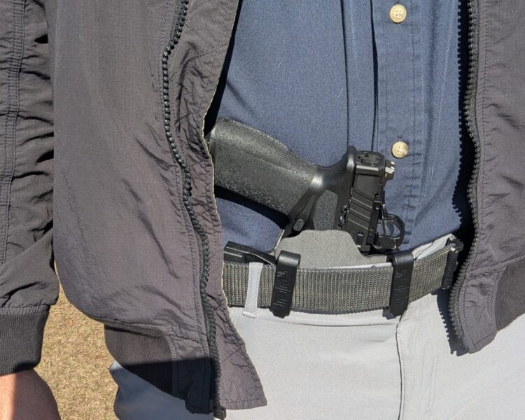 Close-up of a pistol holstered in the appendix position, revealed by an open jacket.