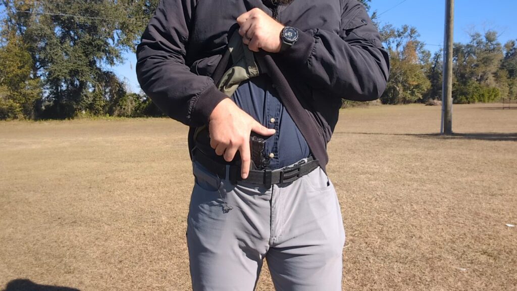 Close-up of a man's hand on the grip of a pistol concealed in an appendix holster, wearing a black jacket and belt.