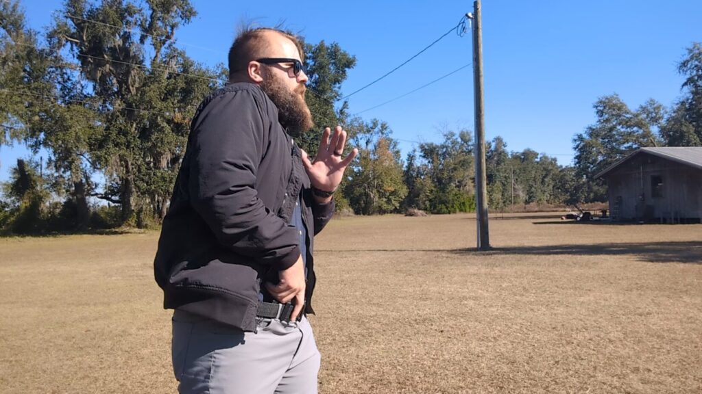 Man in sunglasses and jacket aiming pistol with both hands on an outdoor range.