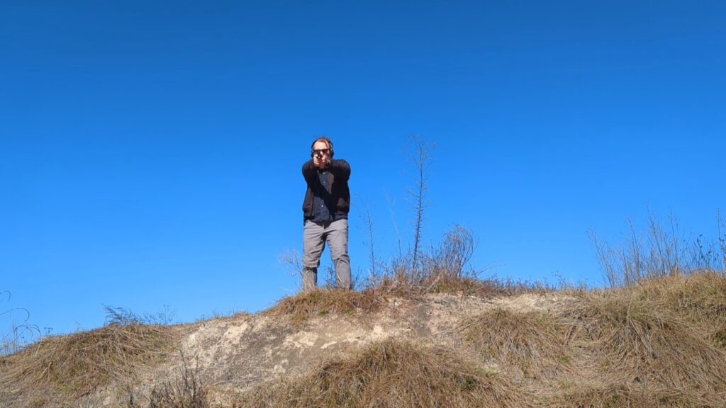 Shooter aiming down from a dirt berm against a blue sky.
