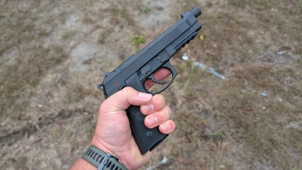 Hand holding a Beretta 92 pistol with a threaded barrel at an outdoor range.