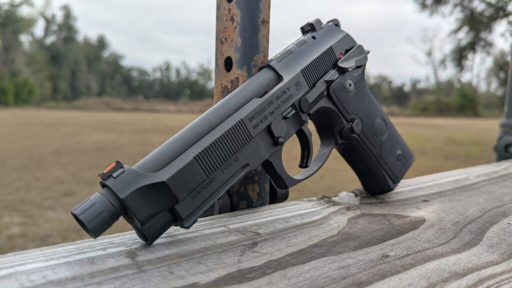 Side profile of a Beretta 92 pistol with a threaded barrel, resting on a wooden rail outdoors.