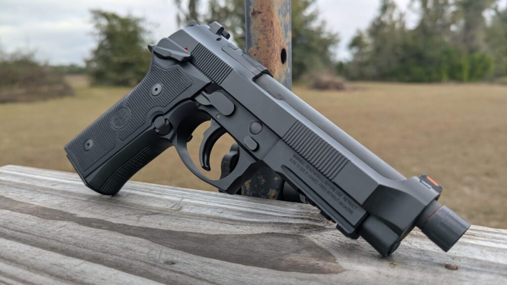 Side profile of a Beretta 92 pistol with a threaded barrel, resting on a wooden rail outdoors.
