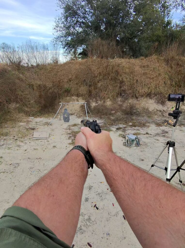 Shooter's point-of-view aiming a Beretta 92 pistol at a steel target stand outdoors.