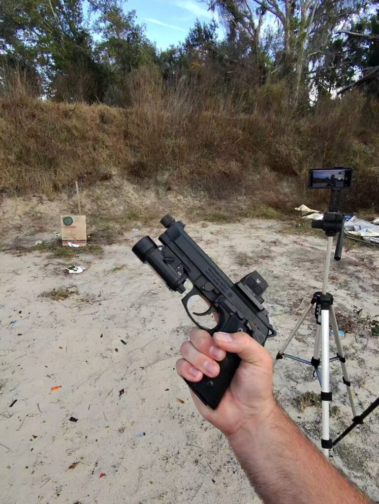 Hand holding a Beretta 92 pistol with a red dot sight and weapon light at an outdoor range.