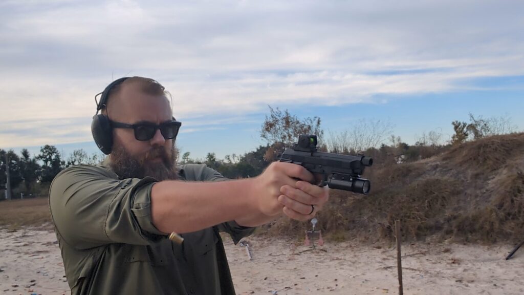 Shooter firing a Beretta 92 pistol with a red dot sight and weapon light at an outdoor range