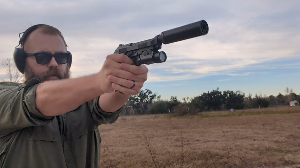 Shooter aiming a suppressed Beretta 92 pistol with a mounted light at an outdoor range.