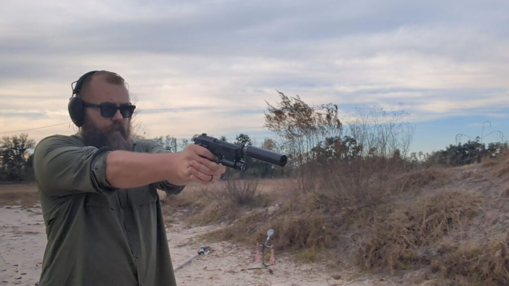 Shooter aiming a suppressed Beretta 92 pistol with a mounted light at an outdoor range.