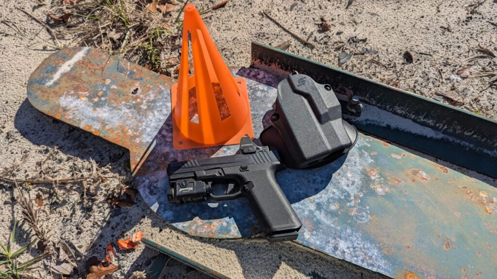 Pistol with red dot sight and light, holster, and cone on the ground.
