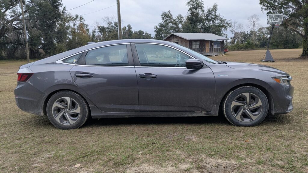 Full side view of a dirty, dark gray sedan (Honda Insight Hybrid) parked on grass.