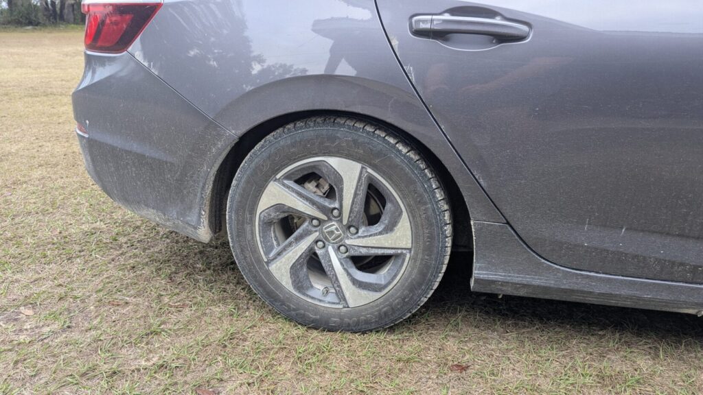 Rear-side view of a dirty, dark gray sedan (Honda) wheel and rear quarter panel in a grassy field.