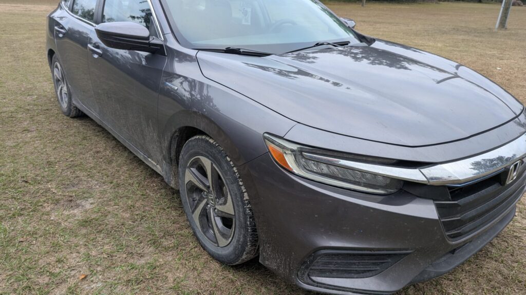 Close-up of the front corner, headlight, and grille of a dirty, dark gray sedan (Honda Insight Hybrid).