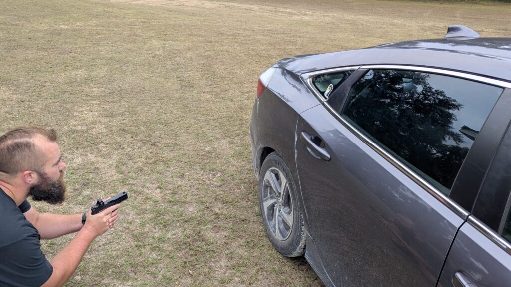 Man aiming a pistol while standing behind the rear door of a dark gray sedan.