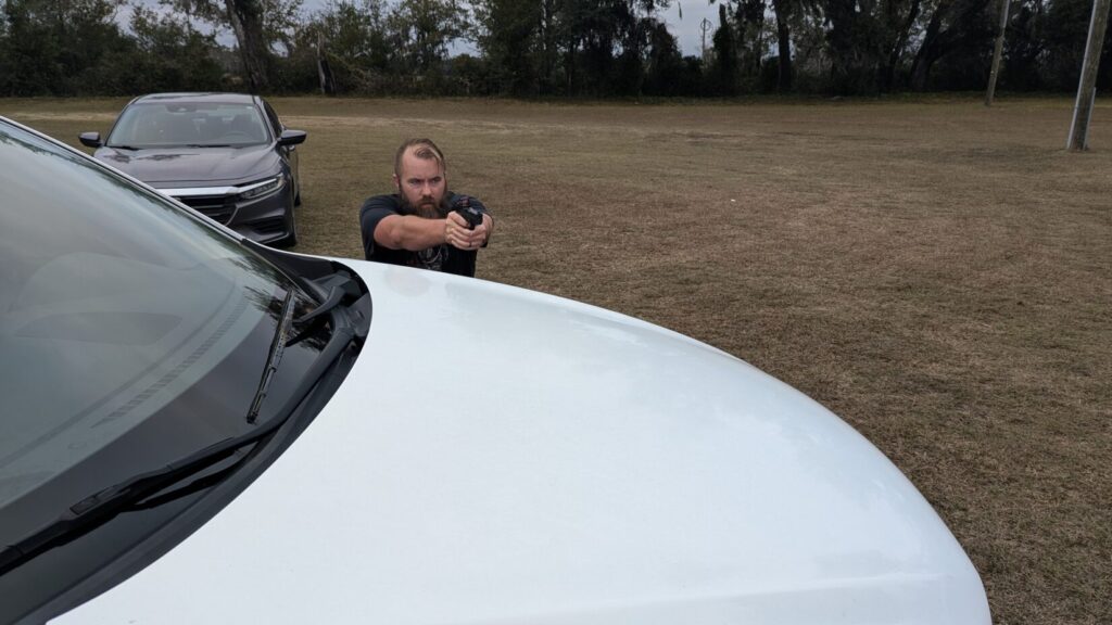 Man aiming a pistol from behind the white hood of a car, using it as cover.